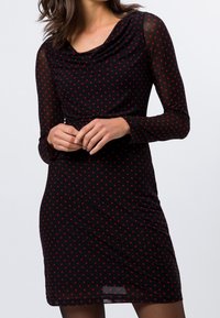 Robe noire avec de longues manches transparentes, décolleté en cowl et pois rouges. Silhouette ajustée avec une taille froncée et une longueur au genou.