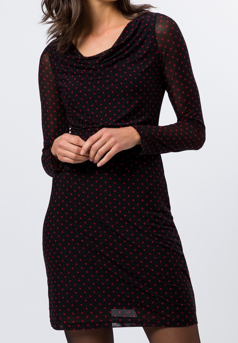 Robe noire avec de longues manches transparentes, décolleté en cowl et pois rouges. Silhouette ajustée avec une taille froncée et une longueur au genou.