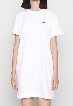 Kvinde iført en løs hvid kortærmet kjole med et lille lilla Adidas-logo på venstre bryst mod en ensfarvet baggrund.