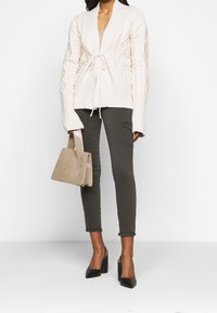 Cárdigan de punto crema con diseño de trenzado, detalle de cordón, combinado con jeans skinny oliva, tacones negros tipo slingback y un bolso beige texturizado.