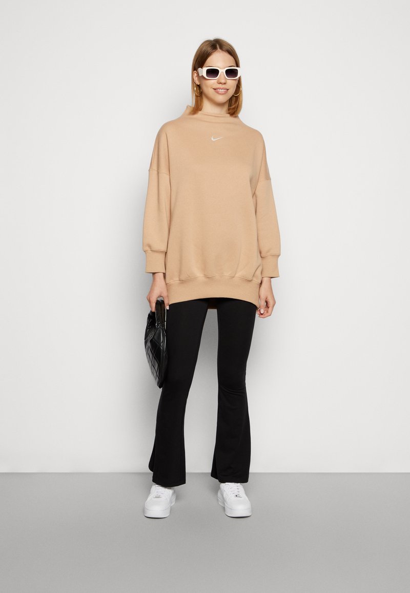 Felpa beige oversize con collo alto, logo Nike bianco. Pantaloni neri svasati, sneakers bianche e piccola borsa nera trapuntata. Occhiali da sole bianchi.