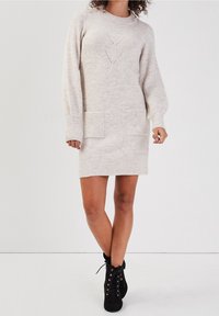 Robe en tricot beige avec des poignets et un ourlet côtelés, dotée de deux poches avant et d'un motif géométrique sur la poitrine. Bottines noires.