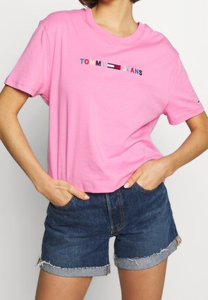 Kobieta ubrana w różową koszulkę Tommy Jeans z wielokolorowym logo oraz ciemne jeansowe szorty z mankietami, dłonie w kieszeniach, na jednolitym tle.