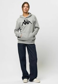 Grijze hoodie met een grote zwarte graphic op de voorkant, voorzien van een trekkoordcapuchon en een kangoeroezak, gecombineerd met donkere denim wijde broek.