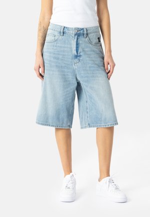 Lyseblå denim Bermuda-shorts båret af en person i en hvid tank top og hvide sneakers, stående på en hvid baggrund.