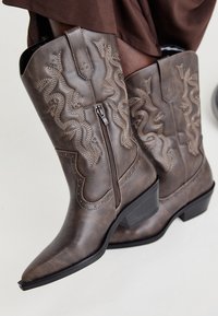 Botas vaqueras de cuero marrón con punta afilada, cremallera lateral y diseño de serpiente texturizado en beige, con tachuelas decorativas a lo largo de los bordes.