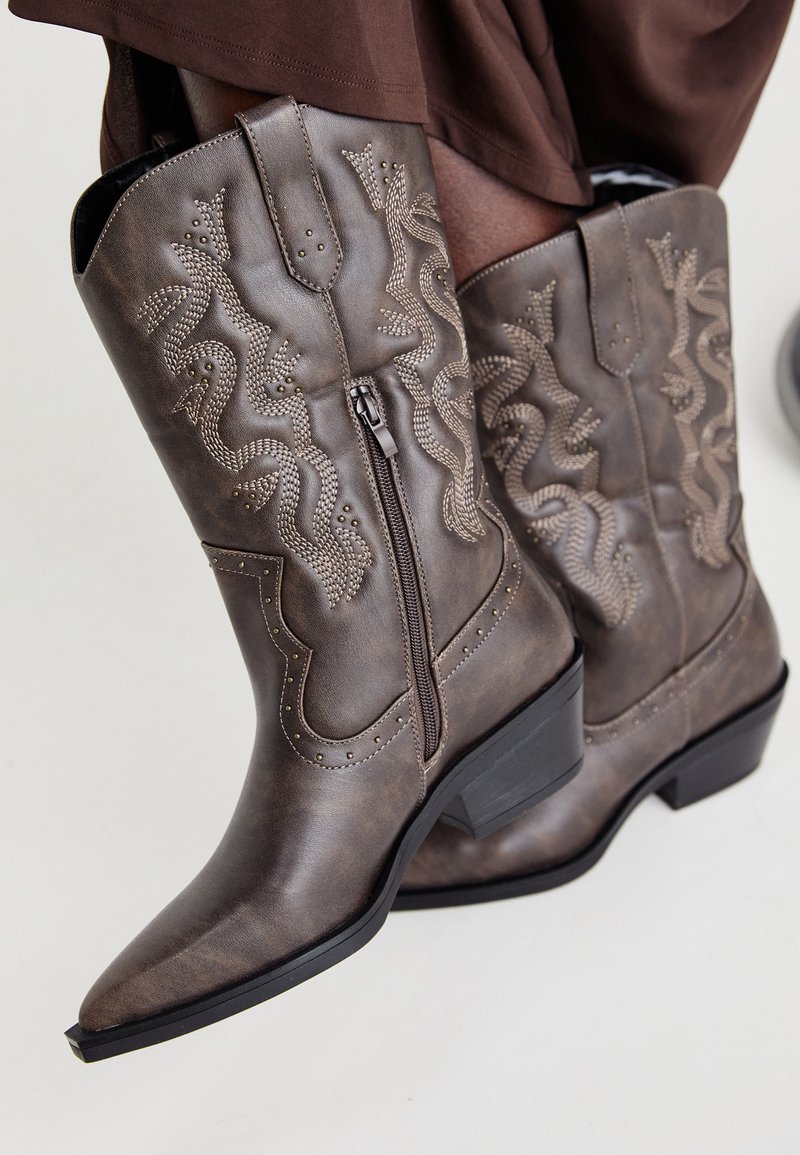 Botas de cowboy em couro castanho com bico pontiagudo, fecho lateral e design texturizado de serpente em bege, apresentando tachas decorativas ao longo das bordas.