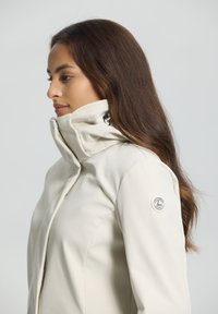 Witte softshell jas met een hoge kraag, voorzien van een zijrits, aansluitend ontwerp en een logo patch op de linker mouw. Soepele textuur.
