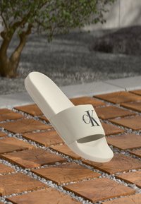 Hvit sandale med stoffstropp som har en svart CK-logo, strukturert s�åle, lett design og jevn finish, plassert på en flislagt bakgrunn.