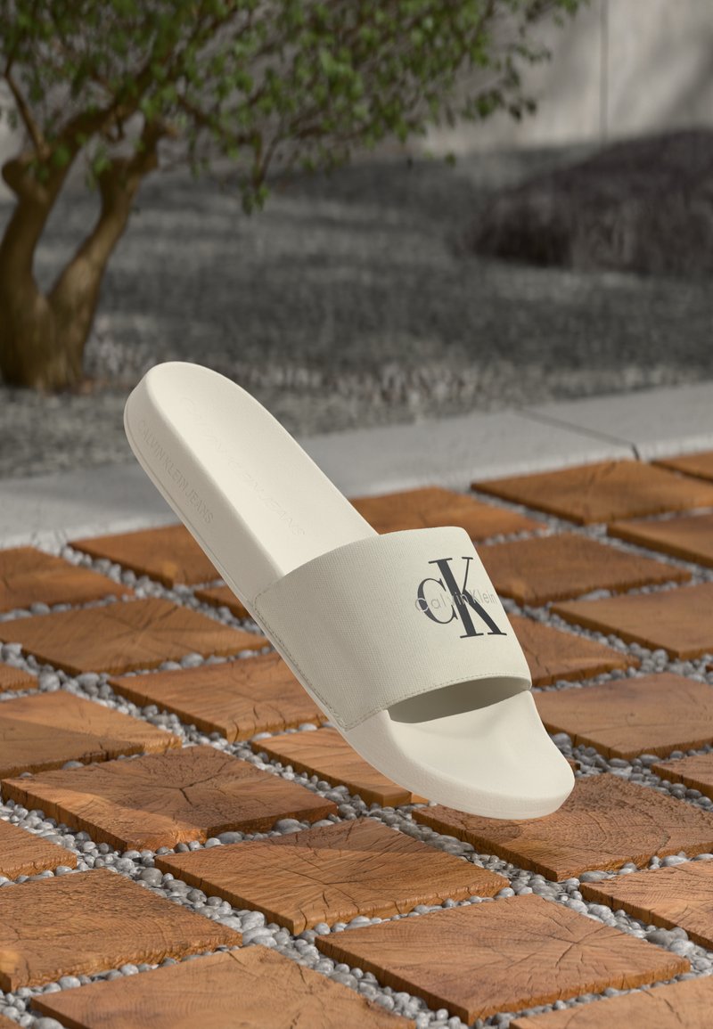 Hvit sandale med stoffstropp som har en svart CK-logo, strukturert såle, lett design og jevn finish, plassert på en flislagt bakgrunn.