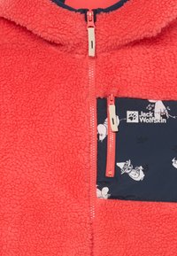 Fleece-takki kirkkaan korallinvärisenä, jossa on tummansiniset taskut kevyellä kuviolla. Edessä vetoketjukiinnitys, jossa on kontrastivärinen vetoketju ja kangastekstuuri.