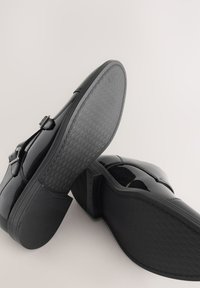 Chaussures habillées en cuir noir brillant avec boucles, montrant une semelle avec un motif texturé détaillé et un talon empilé sur un fond pâle.
