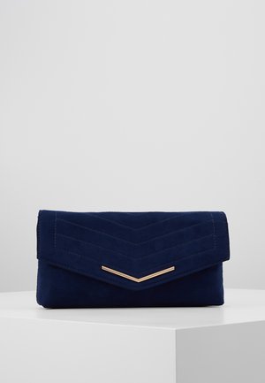 Marineblauwe suède clutch met chevronstiksels en een gouden V-vormige metalen accent op de voorflap, rustend op een wit oppervlak.