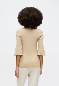 Beige gerippter Stricktop mit lässigen, auslaufenden Ärmeln in Drei-Viertel-Länge, figurbetontem Design und einer glatten Textur. Kombiniert mit hellen, locker sitzenden Hosen.