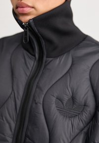 Chaqueta acolchada negra con un cuello alto, que presenta un logo bordado en tono y una textura suave; cierre de cremallera en la parte frontal.