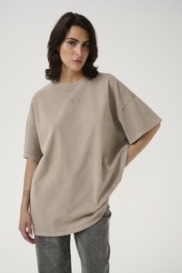 Beige oversized t-shirt with short sleeves, featuring a subtle logo in the center. Fabric has a soft, textured appearance, paired with patterned pants.