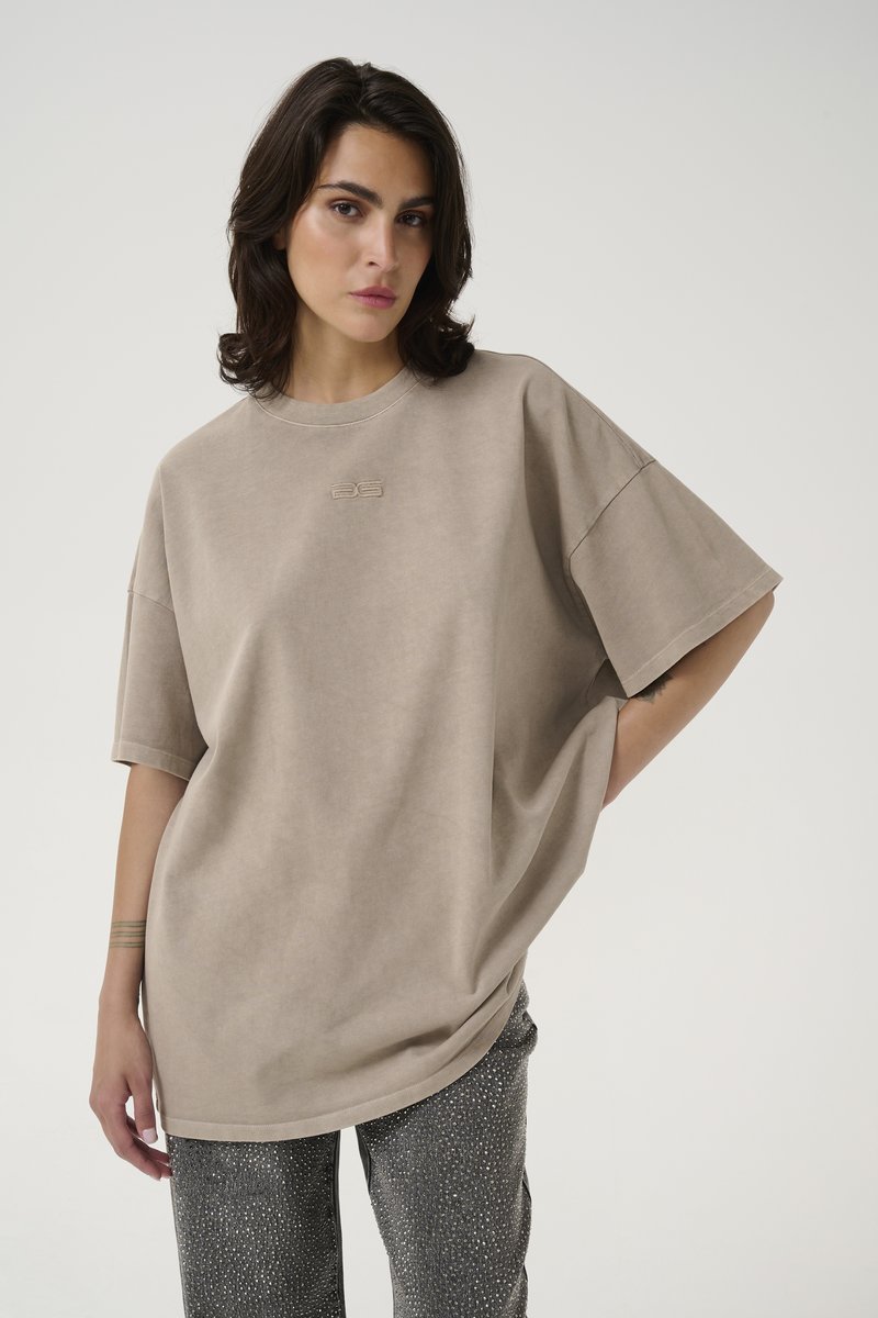 Beige oversized t-shirt with short sleeves, featuring a subtle logo in the center. Fabric has a soft, textured appearance, paired with patterned pants.