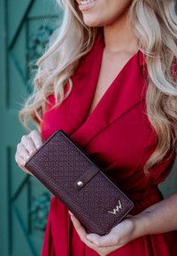 Burgundy wallet with a textured geometric pattern, gold hardware accent, and a button closure, held in a hand against a contrasting background.