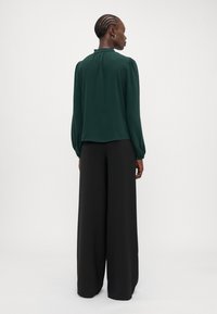 Dark green blouse with puffed sleeves and ruffled neckline, paired with wide black trousers. Smooth fabric, relaxed fit, minimalist design.