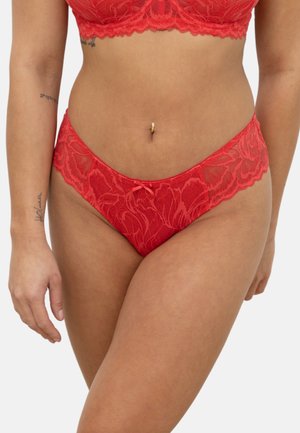 Woman wearing red lace bra and matching panties, showing torso, pierced navel, and arm tattoos against a plain background.