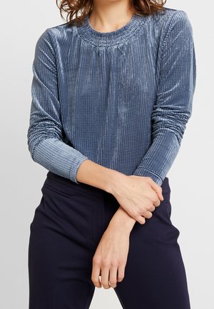 Langärmliges Oberteil aus metallisch blauer Stoff mit einem strukturierten Gittermuster, das einen runden Ausschnitt und geraffte Details an den Schultern aufweist.