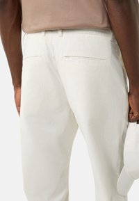 Jasnobeżowe bawełniane spodnie o gładkiej fakturze, z dwoma tylnymi kieszeniami i wygodnym paskiem w talii. W ręku trzymana biała czapka.