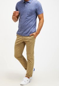 Kortärmad blå polo med en texturerad tyg, beige chinos och vita sneakers med blå detaljer, i en avslappnad ställning.