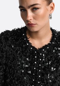 Musta tekstuuriga jakk, millel on karvane disain, kaunistatud sequins'ide ja kivikestega esiava ümber, luues kihilise efekti.