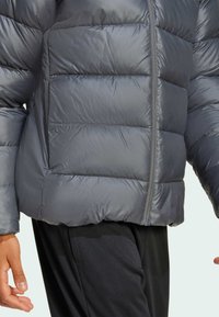 Zbliżenie na osobę w szarej kurtce puchowej z zamkiem, z widoczną ręką, w połączeniu z czarnymi spodniami, na jasnym tle.