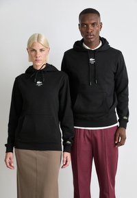Black hoodies made of soft fabric feature kangaroo pockets, drawstring hoods, and embroidered Lacoste logos on the front and sleeves.