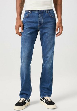 Jeans in denim blu con taglio dritto, dotati di cinque tasche, cuciture a contrasto e chiusura con bottone. Indossati con sneakers nere e bianche.
