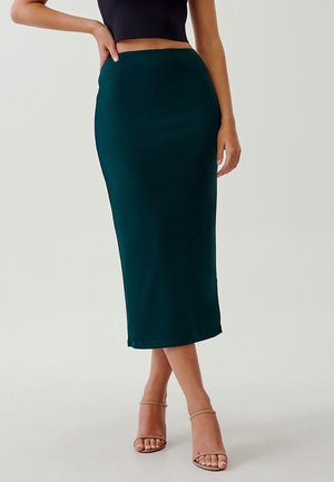 Femme portant une jupe crayon midi taille haute vert foncé, un top court noir et des talons beiges à brides, debout avec une main sur la hanche.