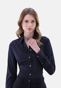 Frau mit blonden Haaren, die ein enges dunkelblaues Hemd mit Punkten und schwarze Hosen trägt, während sie mit einer Hand den Kragen des Hemdes hält.
