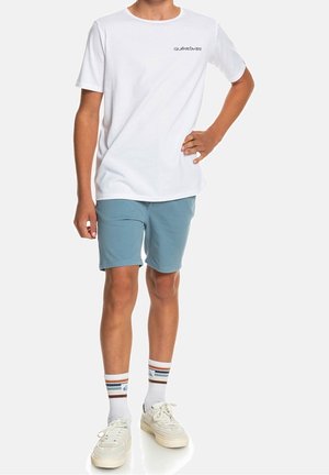 Biały t-shirt z krótkim rękawem z logo Quiksilver, zestawiony z jasnoniebieskimi szortami. Uzupełniony białymi sneakersami i pasującymi, w paski skarpetkami do kolan.