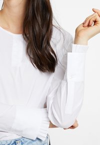 Chemise blanche à manches longues avec des poignets plissés, un col arrondi et des accents de boutons. Le tissu semble lisse et léger.