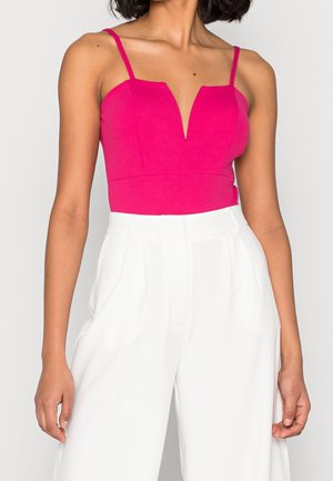 Woman wearing a sleeveless hot pink top with a deep V neckline and high-waisted white wide-leg pants against a plain background.