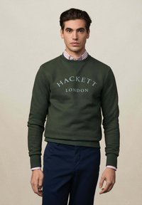 Grüner Kapuzenpullover mit "Hackett London" in hellgrau bedruckt. Rundhalsausschnitt-Design, gerippte Bündchen und Saum. Über einem gestreiften Hemd mit Kragen getragen.