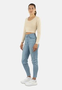 Beige Langarm-Crop-Top kombiniert mit hochgeschnürten hellblauen Jeans und weißen Sneakern. Das Top hat einen runden Halsausschnitt und eine figurbetonte Passform.