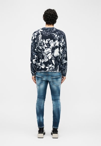 Homme aux cheveux bouclés portant un pull à fleurs bleu marine et blanc, un jean bleu délavé et des bottes noires, debout de dos sur un fond blanc.