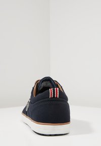 Navy canvas sneaker featuring a brown leather accent at the heel, white sole, and a striped red and white tab at the back.