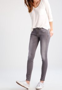 Šedé skinny džíny s nízkým pasem, detailním šitím a jemně ošuntělým vzhledem. V kombinaci s svetrem v světlé barvě a bílými teniskami.