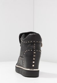 Zapatilla deportiva con cuña de cuero negro decorada con tachuelas y cremalleras plateadas, con puntera redondeada, parte superior texturizada y suela de goma plana.