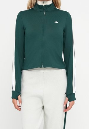 Osoba w ciemnozielonej kurtce zapinanej na zamek z białymi paskami na rękawach i jasnoszarych spodniach z czarnym logo przy kieszeni.