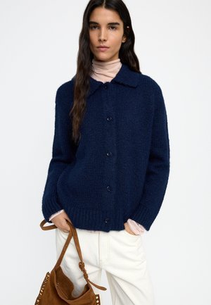 Kobieta ubrana w granatowy sweter na guziki z jasnoróżowym golfem, białe spodnie, trzymająca brązową zamszową torebkę z ćwiekami.