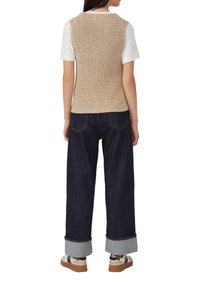 Vrouw draagt een beige gebreide vest over een wit T-shirt met korte mouwen, donkere wijde, opgerolde jeans en witte sneakers met rubberen zolen, staand met haar rug naar je toe.