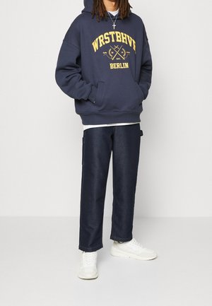 Persona con felpa con cappuccio blu navy con scritta gialla "WRSTBHVE Berlin" e jeans scuri, in piedi con le mani nelle tasche della felpa, scarpe da ginnastica bianche visibili.