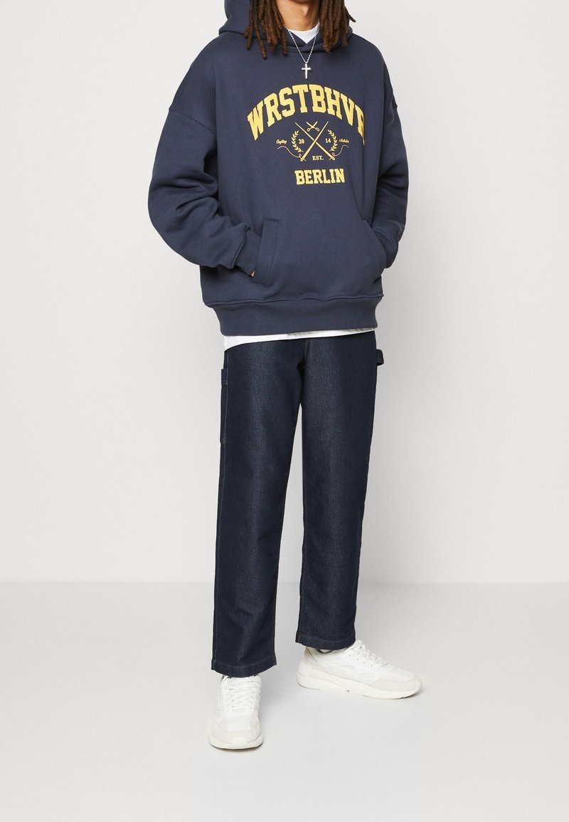 Personne portant un sweat à capuche bleu marine avec le texte jaune "WRSTBHVE Berlin" et un jean foncé, debout les mains dans la poche du sweat, baskets blanches visibles.