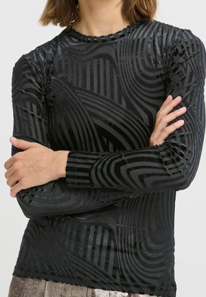 Personne portant un haut noir à manches longues avec des motifs en velours brillants géométriques et rayés, bras croisés sur la poitrine.