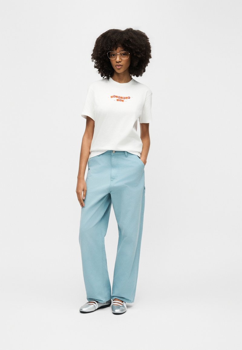 Femme aux cheveux bouclés et portant des lunettes, vêtue d'un t-shirt blanc à motif, d'un pantalon ample bleu clair et de ballerines argentées, debout avec une main dans la poche.