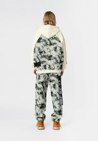 Sudadera con capucha en color crema con un patrón texturizado en verde y blanco en la parte trasera, combinada con pantalones a juego con el mismo patrón y zapatos color marrón claro.